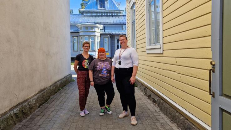 Kolme hymyilevää ihmistä seisovat vierekkäin kahden rakennuksen välissä ulkona. Tuula Enbuska, Reetta Vähä ja Liisa Kurtti.
